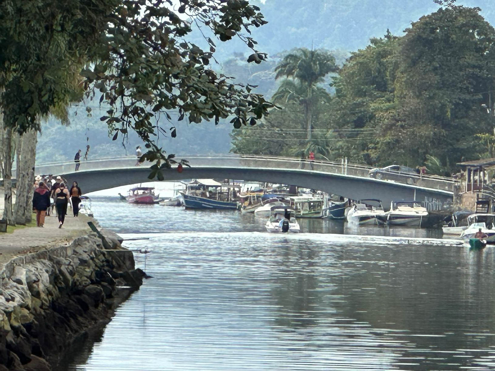 Canal com barcos atracados, atravessado por uma ponte, com pessoas a caminhar na margem e vegetação densa ao fundo.