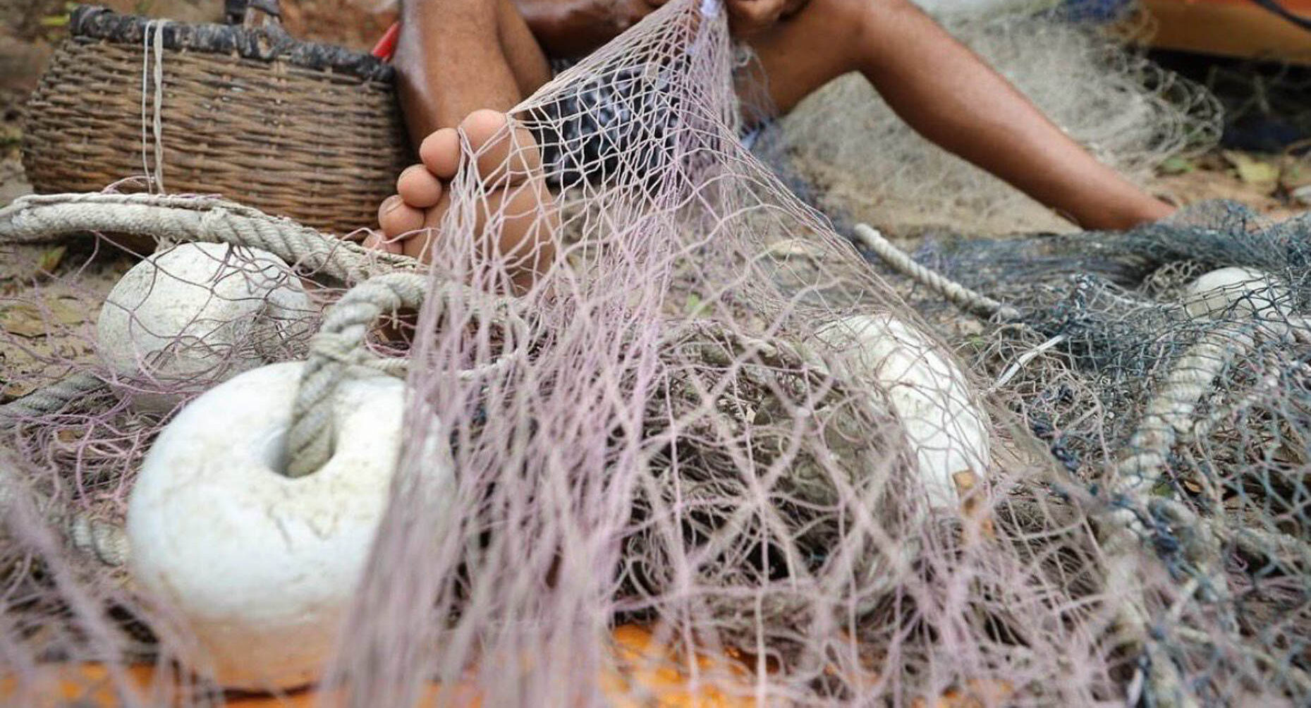 Pescador a remendar uma rede de pesca, com boias e um cesto de vime à volta.